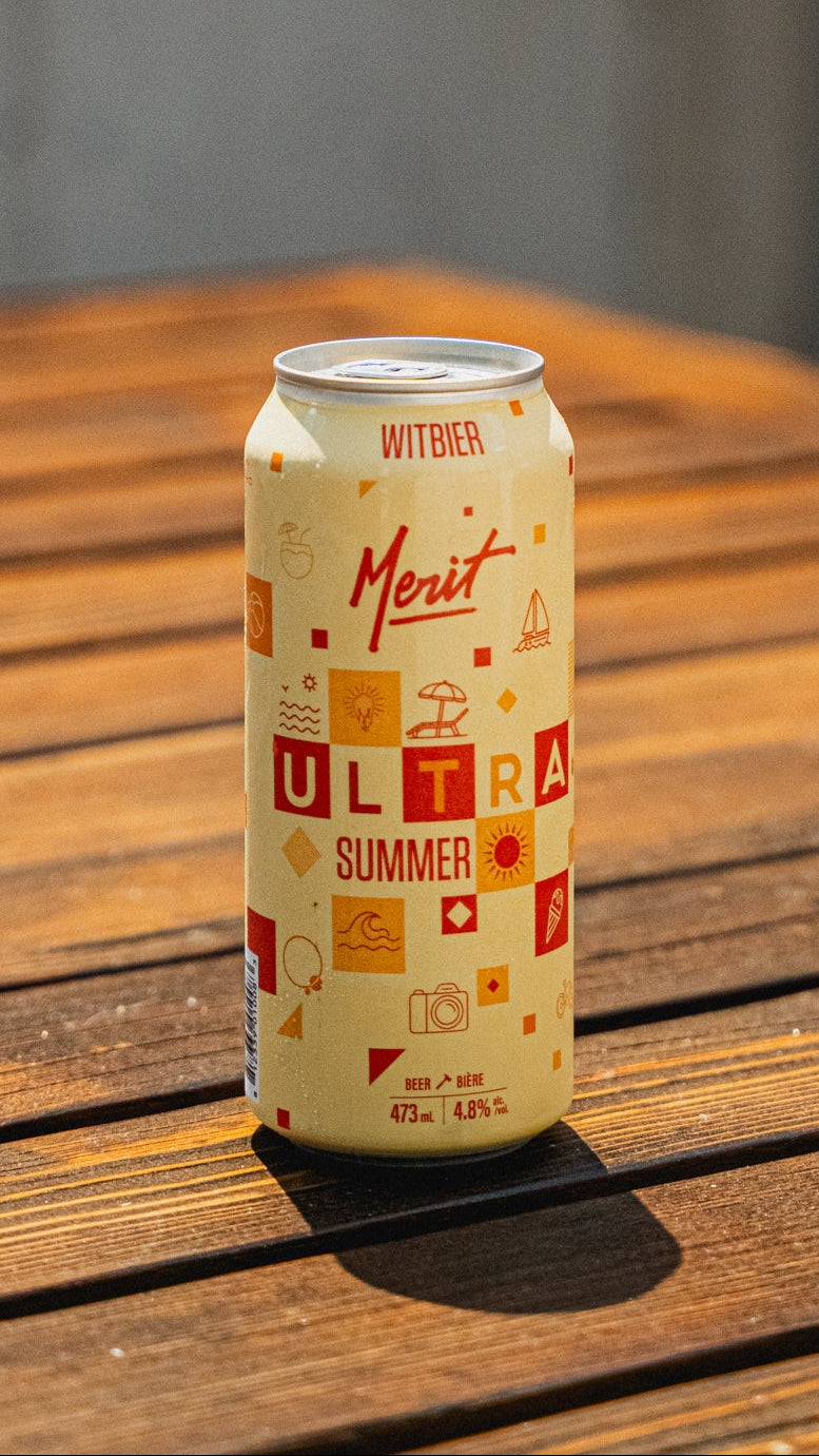 A single can of Merit Ultra Summer Witberger non-alcoholic beer stands on a wooden table in warm natural sunlight, with a blurred gray background.